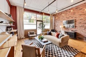 Beige couch on a graphic black and white rug. Large, factory windows and exposed brick frame the room.