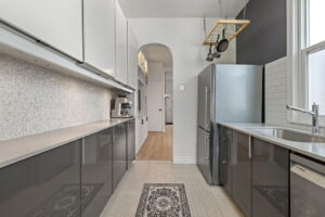Galley kitchen at 21 Blackburn St featuring glossy lower cabinets, white upper cabinetry, subway tile backsplash, stainless steel appliances, and an arched doorway to the living/dining room.