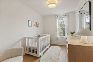Bright second-floor bedroom at 21 Blackburn St styled as a nursery, with a white crib, light wood dresser, soft neutral décor, and two windows facing the backyard.