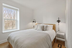 Second-floor bedroom at 21 Blackburn St with a queen bed, soft neutral bedding, black wall-mounted reading lights, and a large window overlooking mature trees.