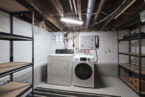 Basement laundry area at 21 Blackburn St with washer and dryer, exposed beams and ducts, concrete floors, and open shelving for storage.