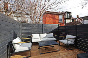 Backyard seating area at 21 Blackburn St with modern outdoor furniture on a wood deck, enclosed by black privacy fencing and surrounded by mature trees and neighbouring homes.