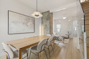 Open dining and living area at 21 Blackburn St, featuring a light wood table, modern pendant lighting, soft neutral furnishings, and a partial concrete accent wall.