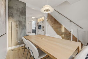Dining area at 21 Blackburn St with a light wood table, modern pendant light, concrete accent wall, and glass-railed staircase leading to the upper level.