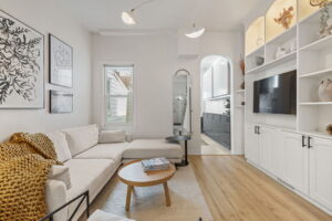 Living room at 21 Blackburn St with a white sectional sofa, illuminated built-in shelving around the TV, and an arched opening leading to the updated kitchen.