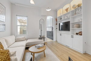 Living room at 21 Blackburn St with a white sectional sofa, illuminated built-in shelving around the TV, and an arched opening leading to the updated kitchen.