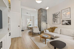 View from the living room at 21 Blackburn St toward the dining area, showing the white sectional sofa, modern artwork, concrete accent column, and light-filled front window.